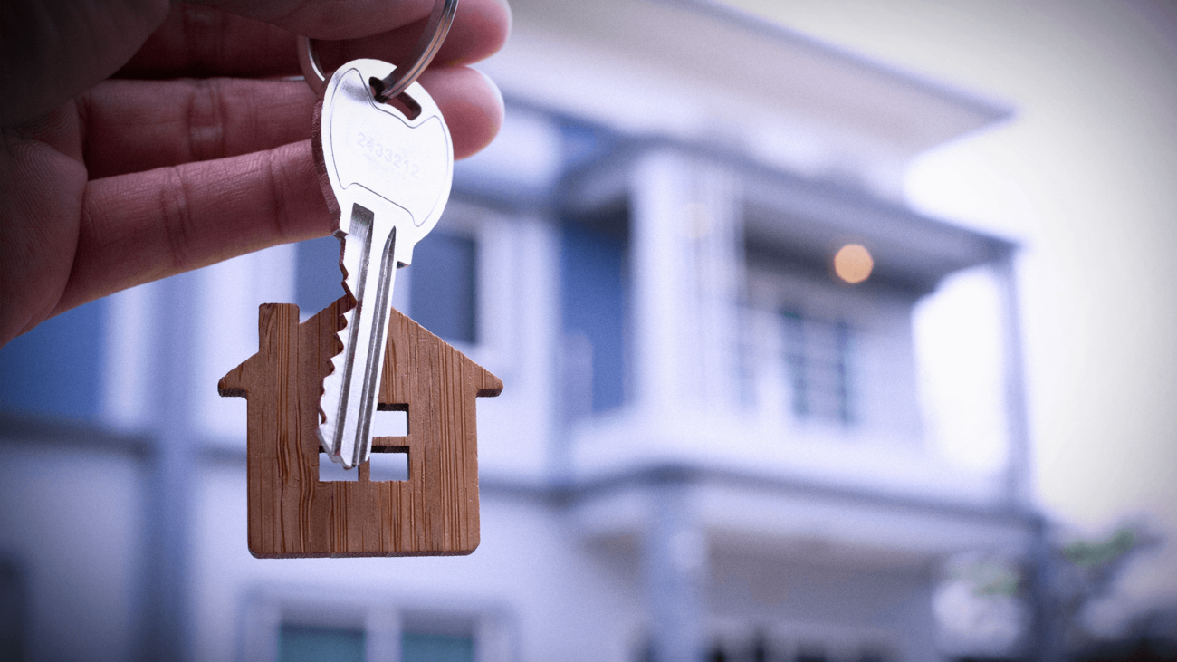 A person is holding a key with a house keychain in front of a house.