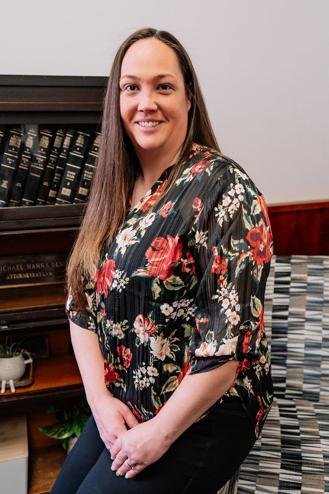 A woman in a floral shirt is sitting on a stool in front of a bookshelf.