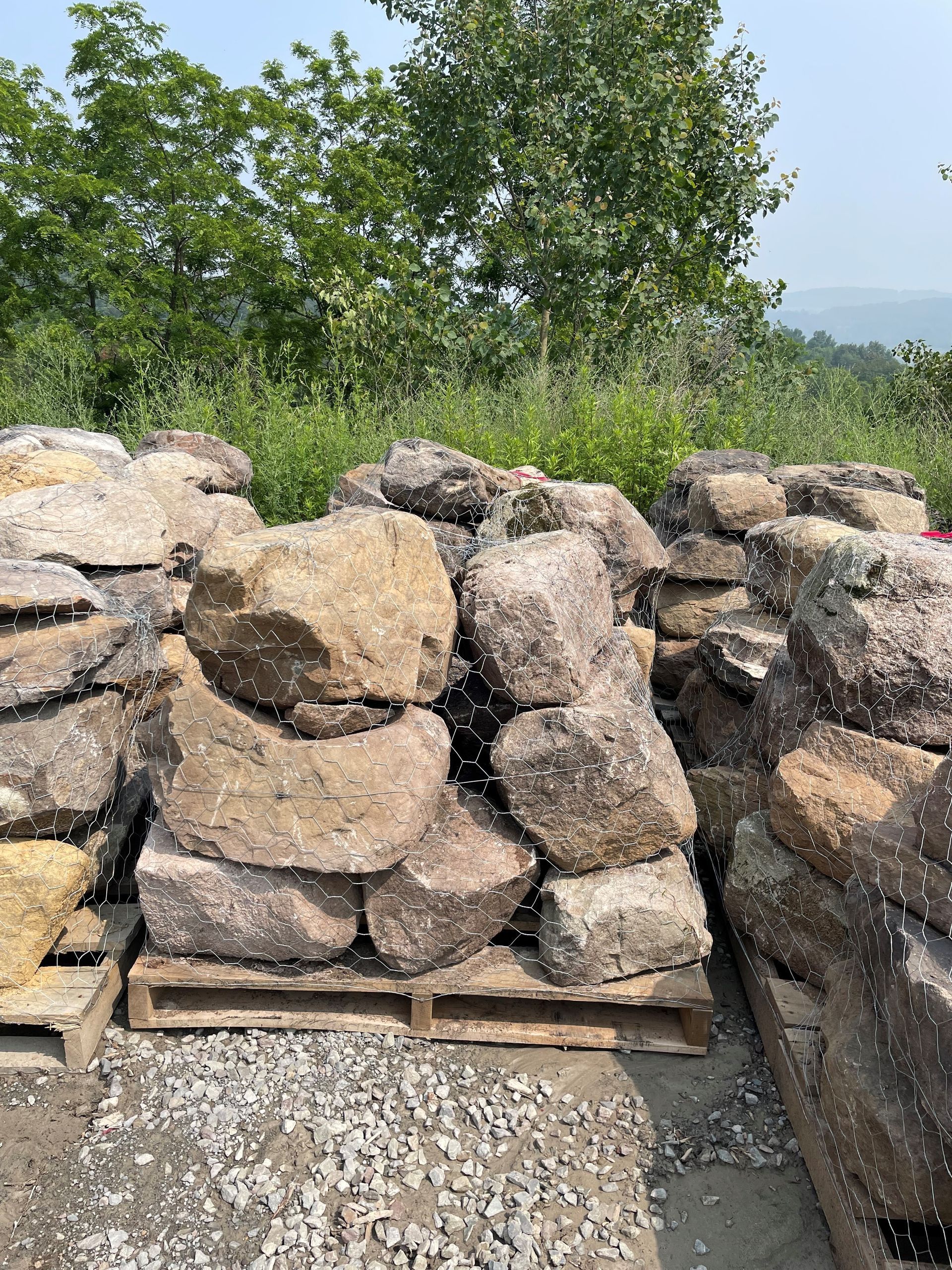 Large brown landscape boulders stacked on wooden pallets in an outdoor yard with greenery in the background.