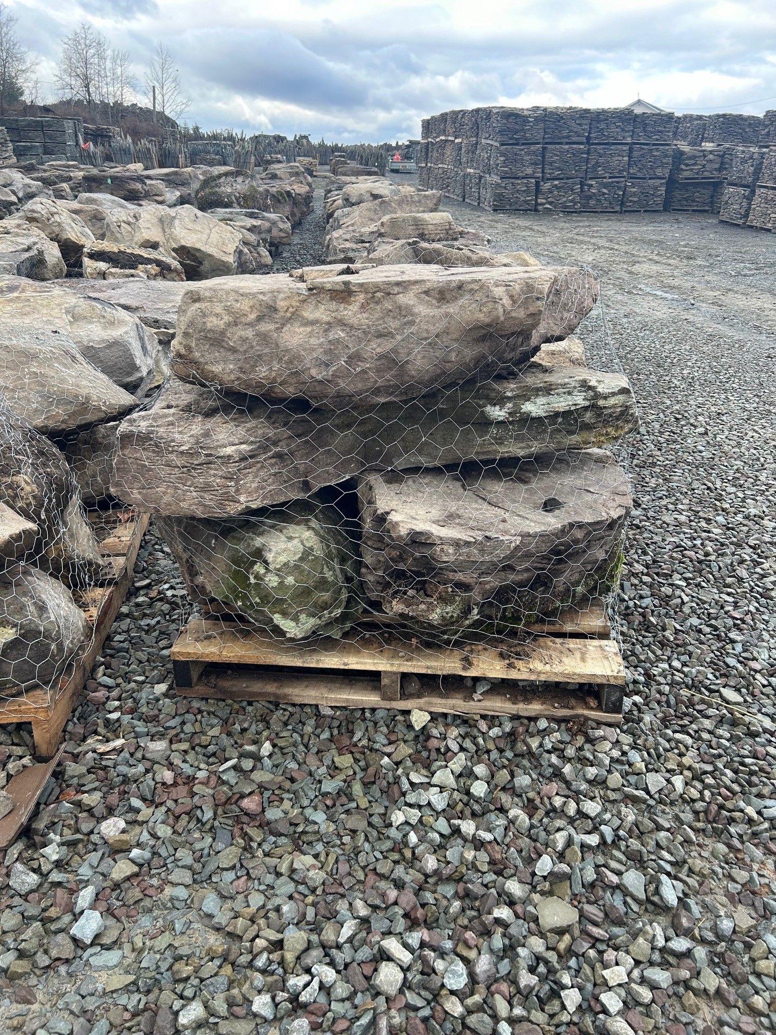 Stacked large, rough, natural stone slabs sitting on a wooden pallet in a gravel yard under an overcast sky.