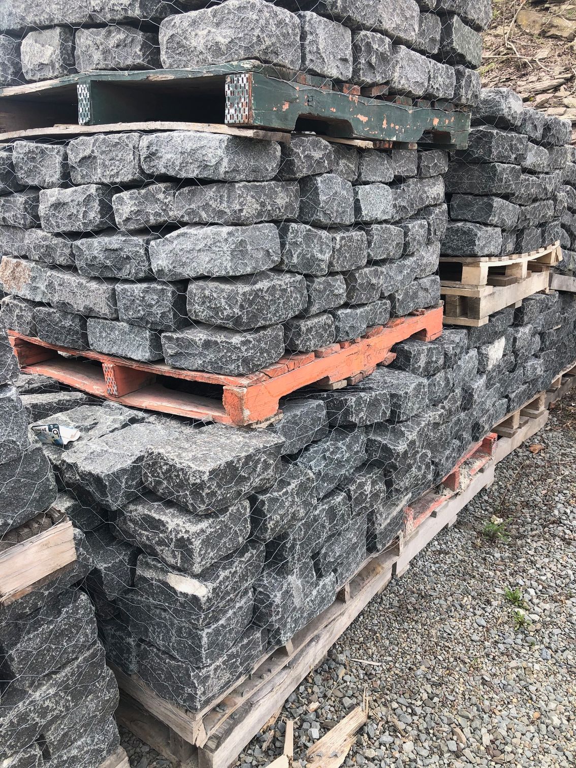 Stacked pallets of dark grey rectangular stone pavers resting on a gravel surface.