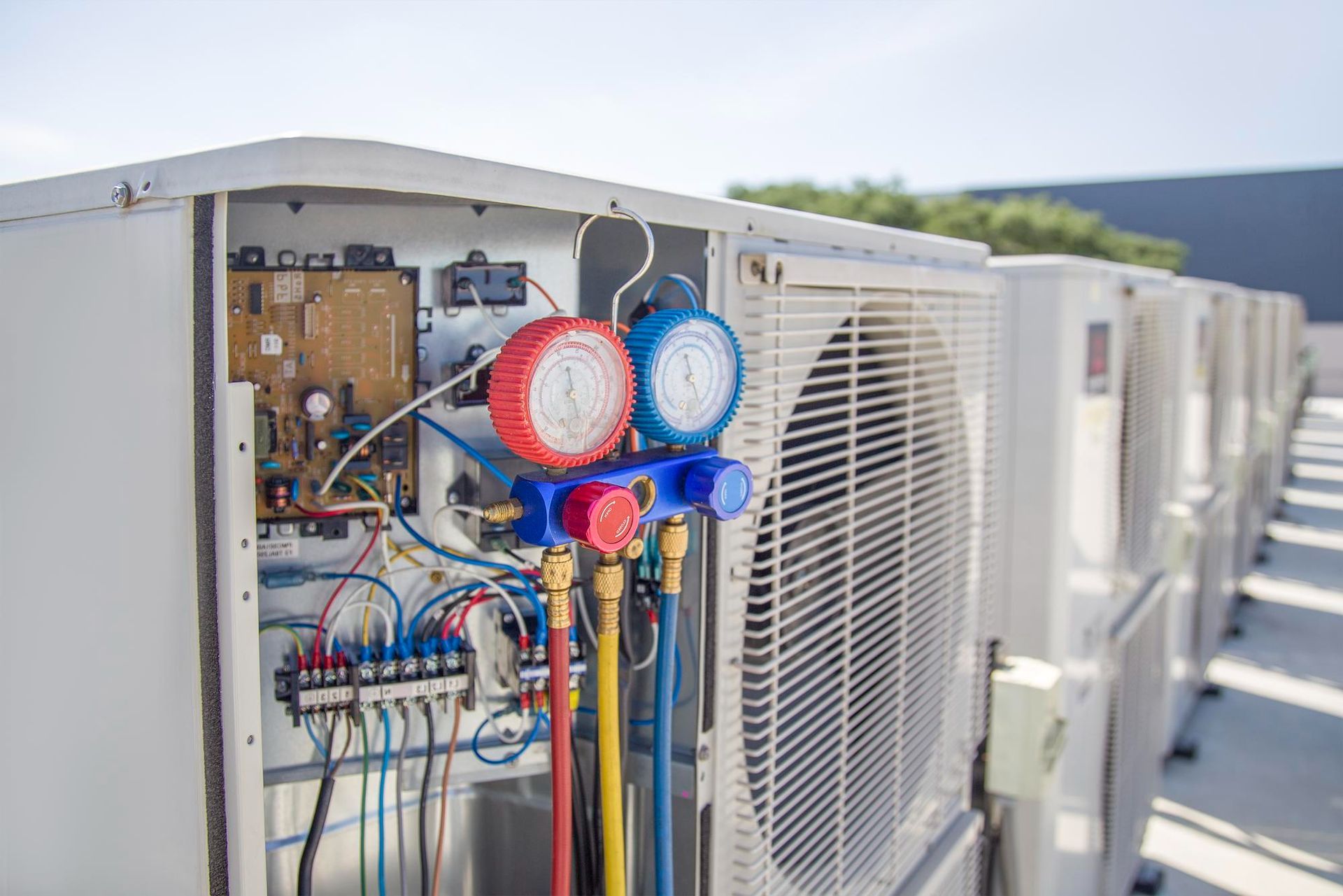 AC unit being serviced with gauges, wires, and tubes connected; row of similar units in background.
