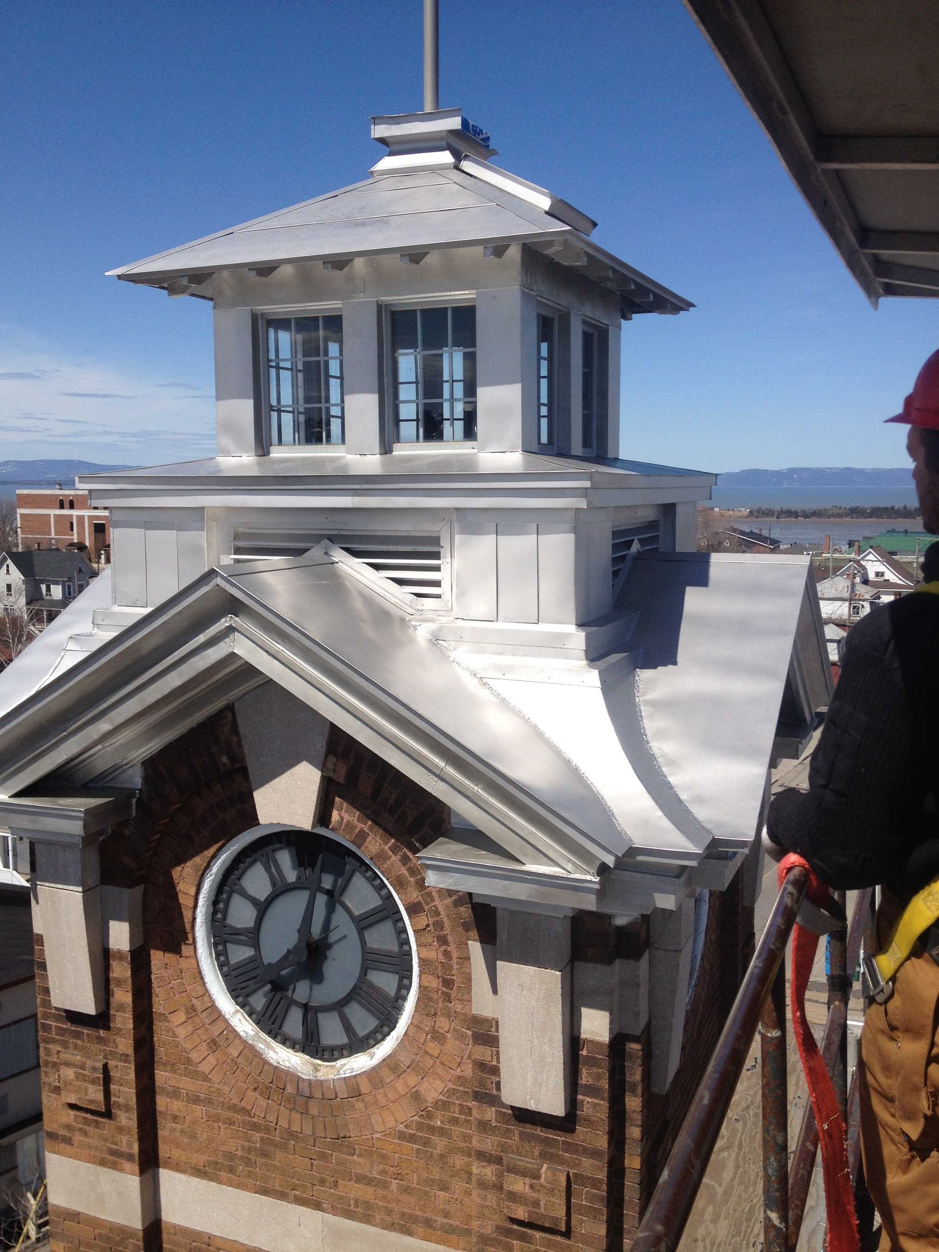 Clock tower with silver roof, brick facade, and workman on a lift. Sunny day.