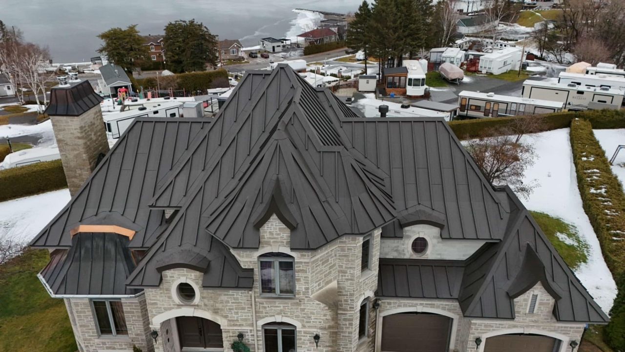 Aerial view of a gray roofed house with stonework, a snow-covered yard, and a lake in the background.