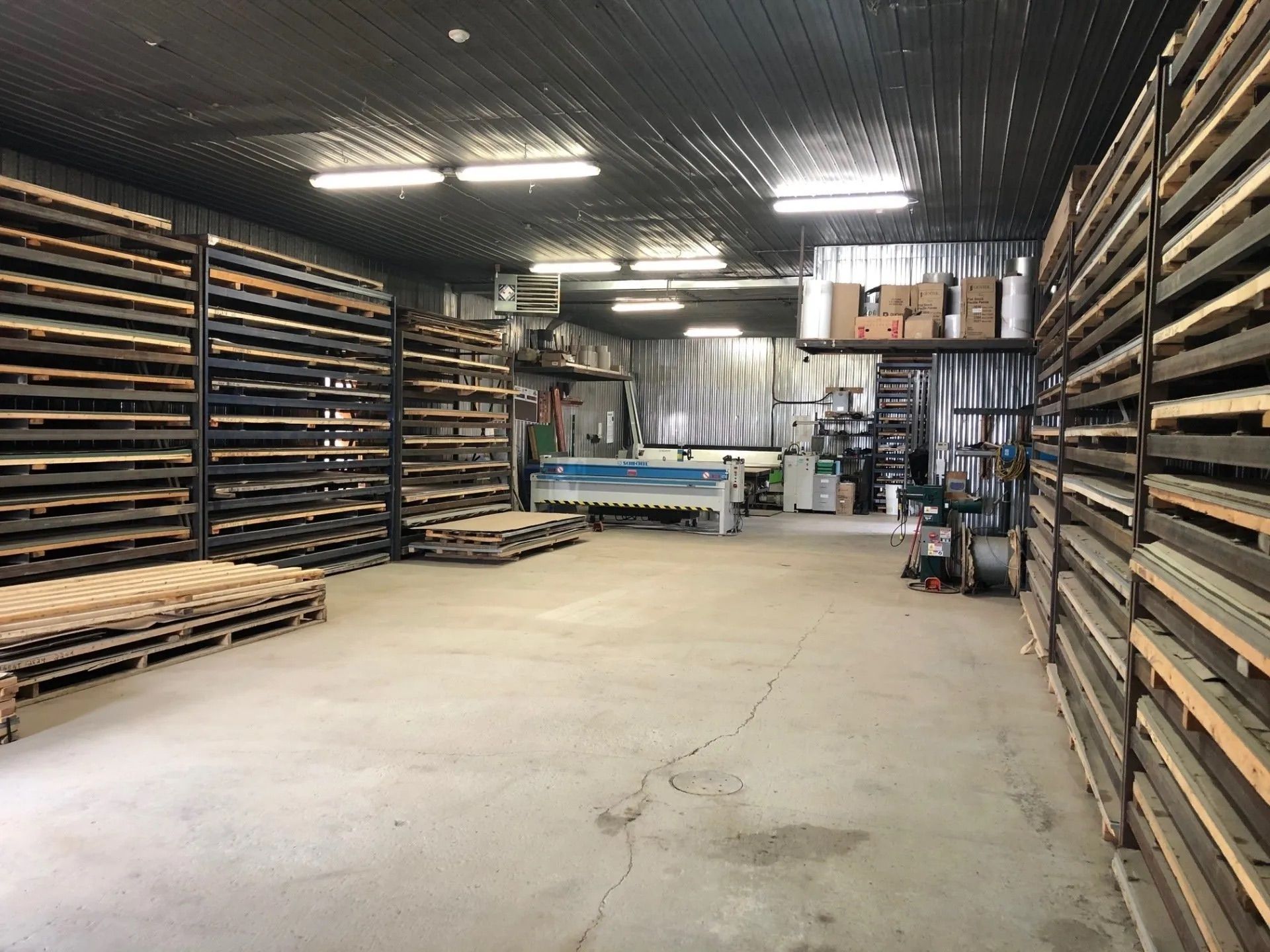 Warehouse interior with metal shelving filled with sheets of material. Pallets and machinery are also visible.