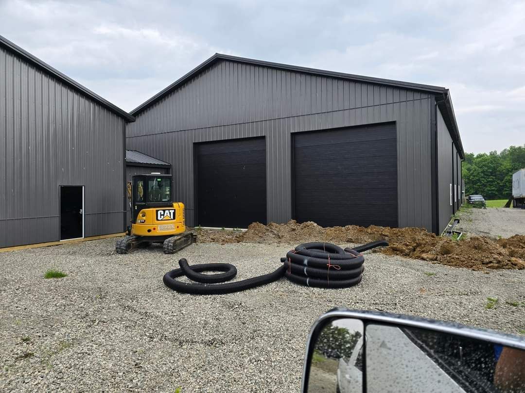 Small excavator near dark metal buildings with black garage doors; black hose in foreground.