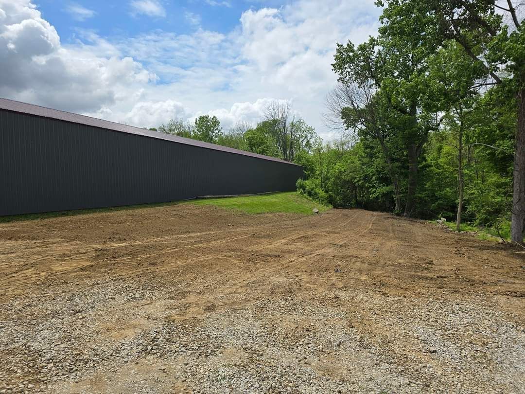 Dirt field in front of a gray building and trees under a cloudy sky.