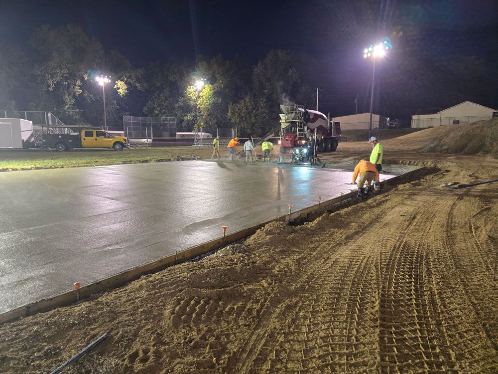 Construction workers pouring concrete at night. A cement truck is visible, with lights illuminating the work.