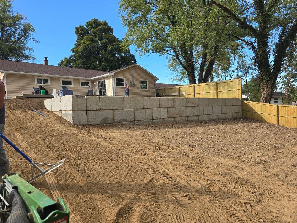 Yard with dirt, retaining wall made of concrete blocks, and house under a blue sky.