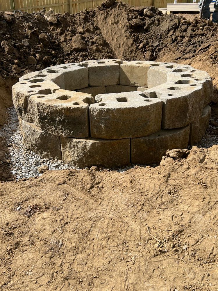 Stone fire pit under construction in a dirt pit. Gravel base and surrounding soil visible.