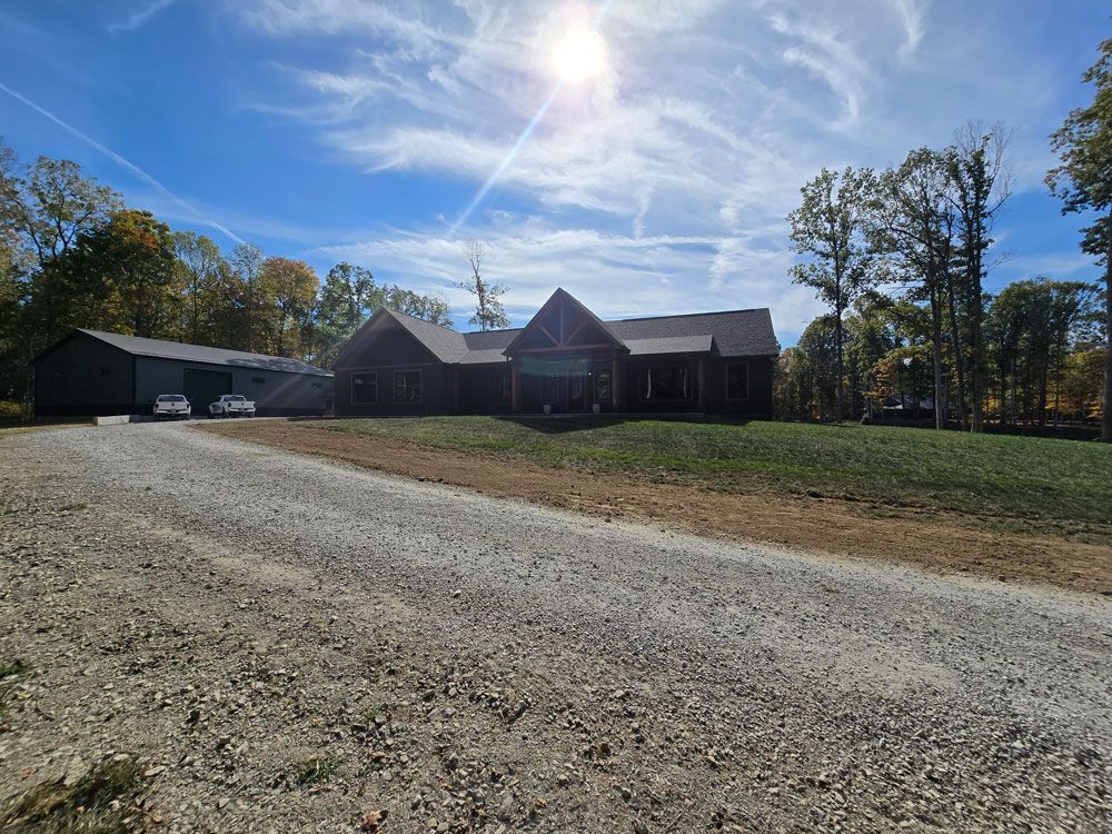 Gravel driveway leads to a dark-colored house with attached garage under a bright sun.