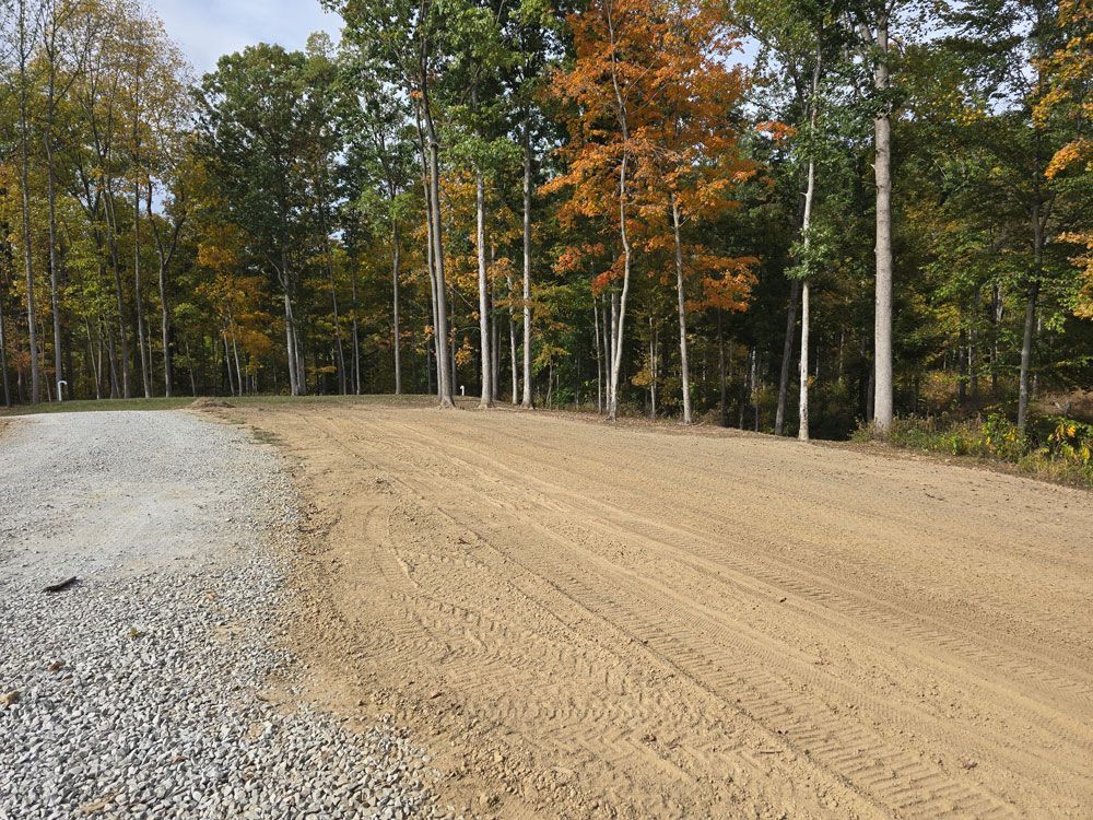 A cleared, sandy area next to a gravel driveway, with a forest backdrop of trees with fall foliage.