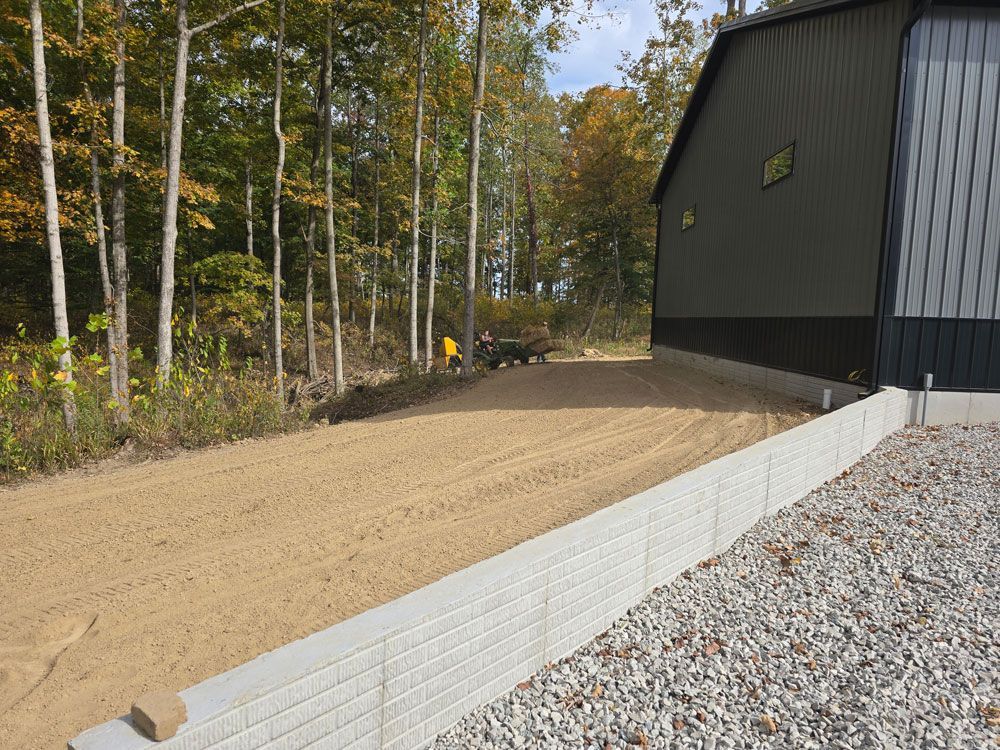 A gravel driveway next to a retaining wall and a dark building; trees are in the background.