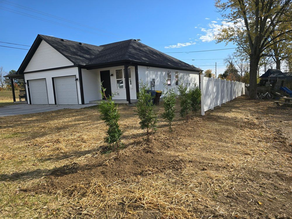White house with black trim, garage doors, and a white fence on a sunny day.