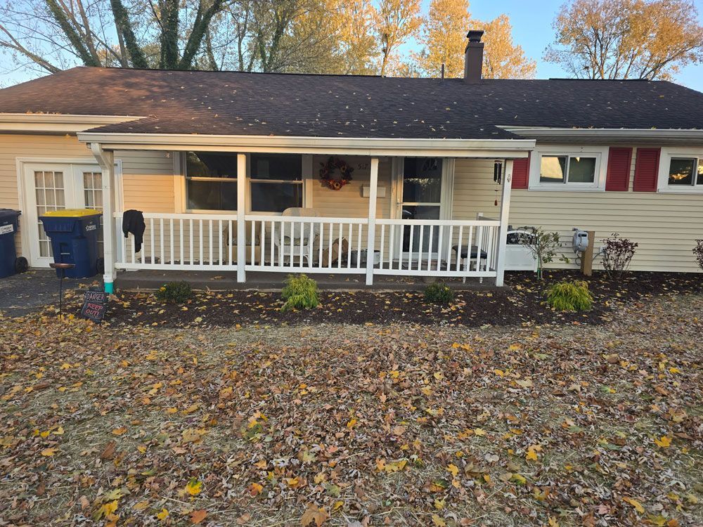 A one-story beige house with a porch and white railing, brown roof, and fall leaves on the ground.