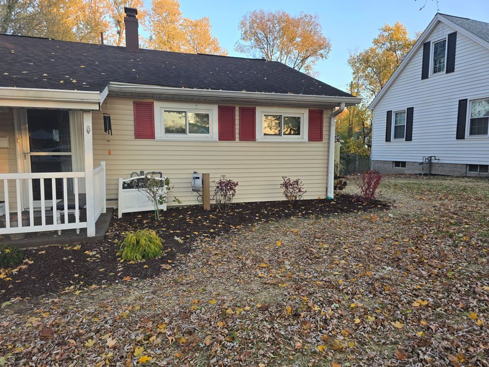 Tan house with red shutters and flower bed; autumn leaves cover the lawn.