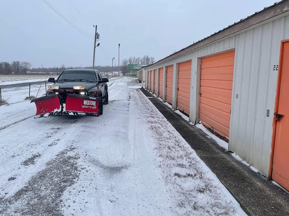 Truck with snowplow clearing snow from a storage unit driveway. Orange doors line the right side.