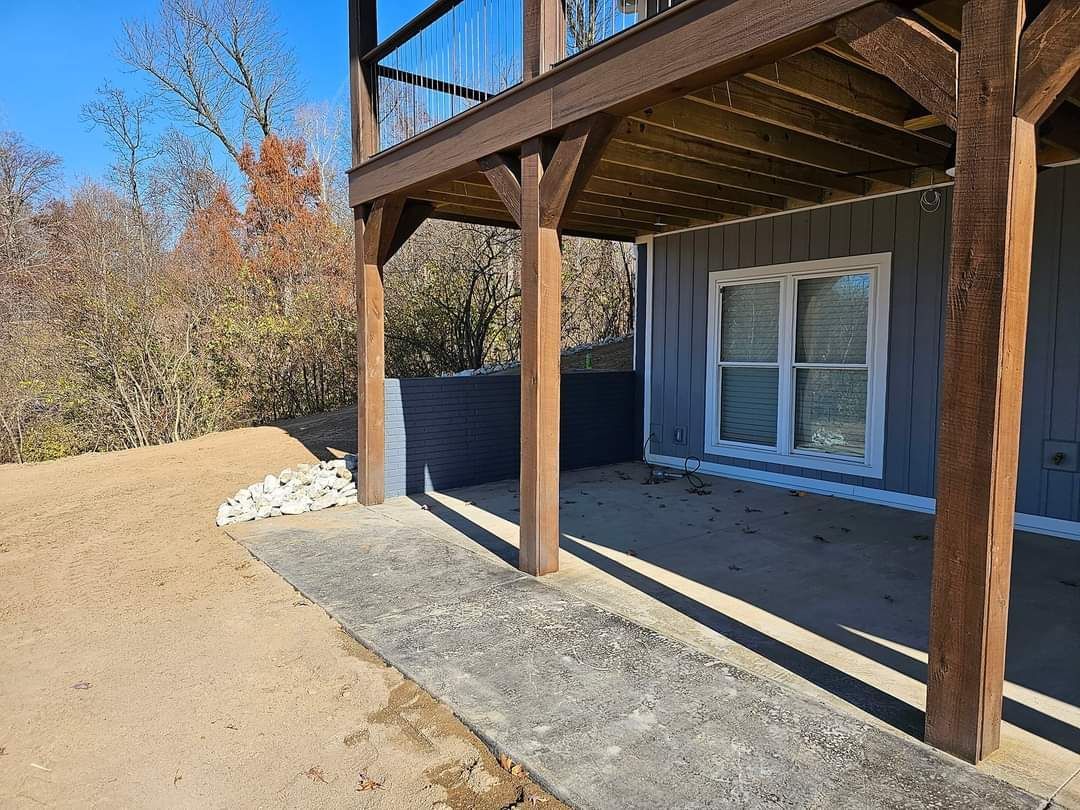 A gray patio beneath a wooden deck, next to a building with a white-framed window.