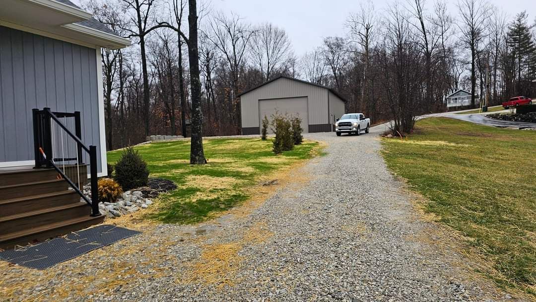 Gravel driveway leads to a garage; a white pickup truck is driving up. A house is on the left, trees in the background.