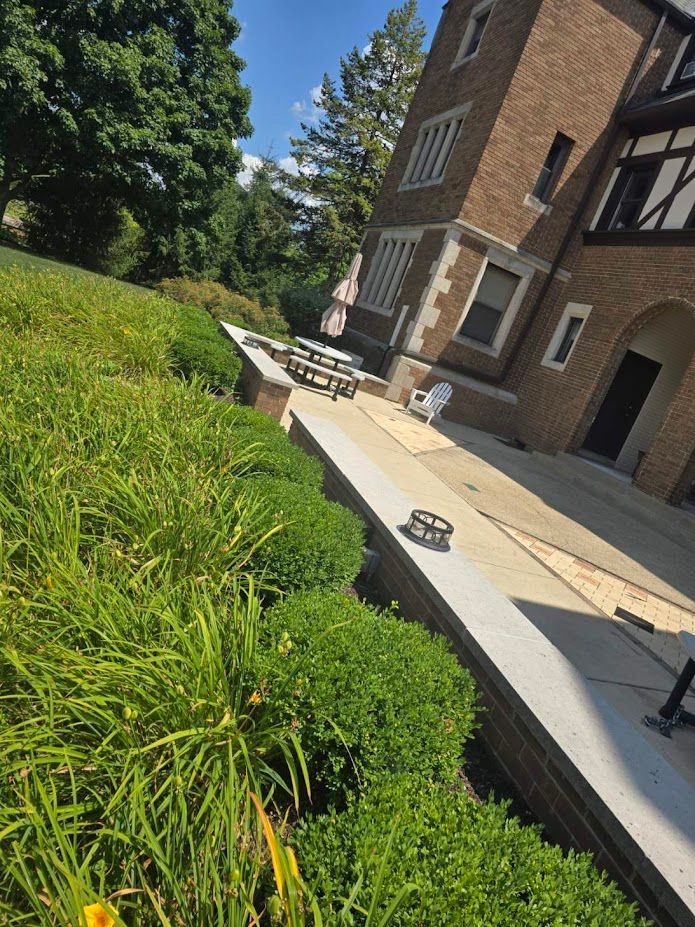 Brick building with stone patio and green shrubbery. Blue sky in background.
