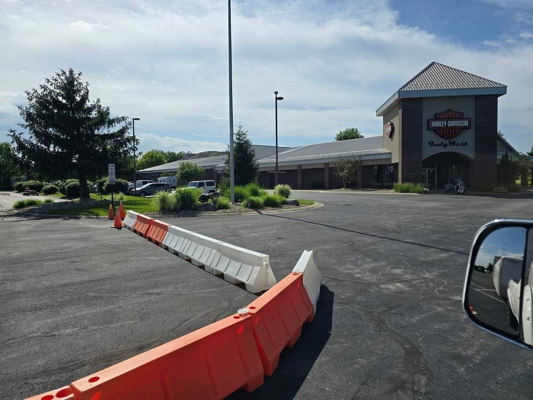 Orange and white barriers block a parking lot entrance near a Harley-Davidson dealership on a sunny day.
