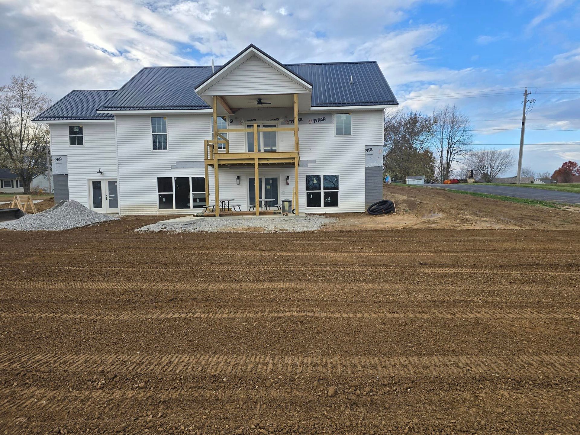 Two-story house under construction with a deck, on a brown field under a cloudy sky.