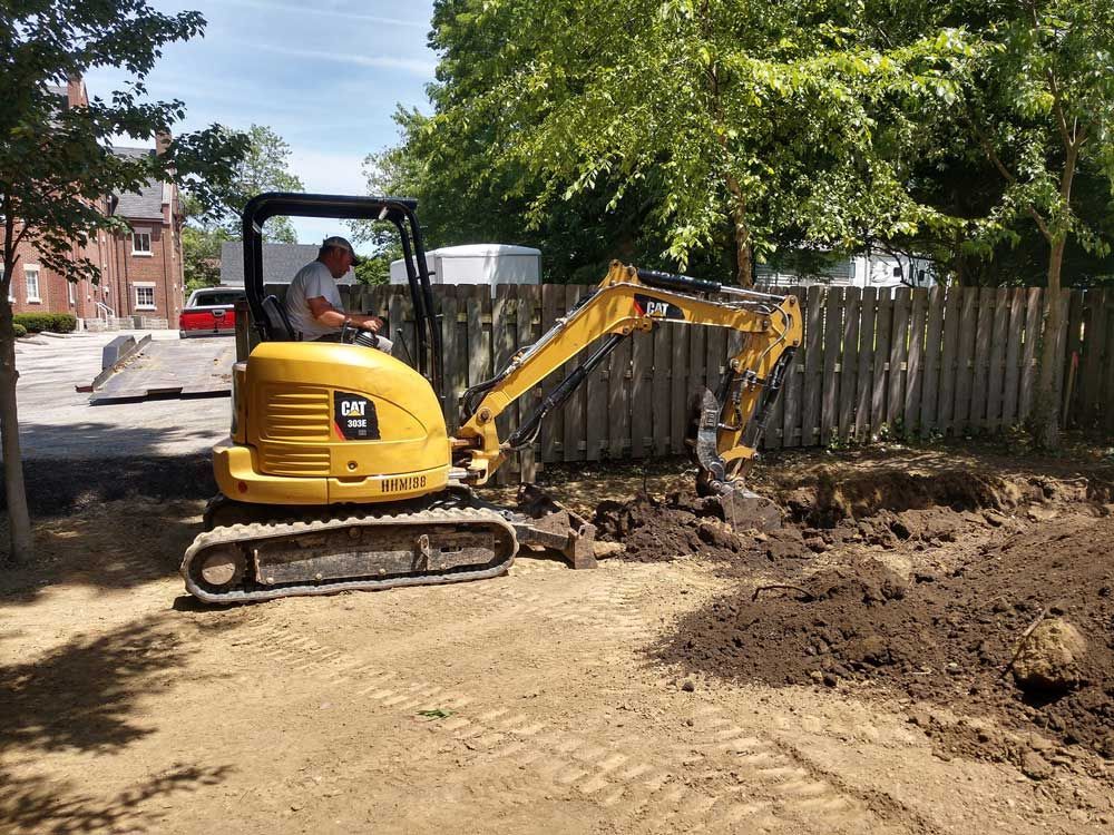 Man operating a yellow excavator digging in a dirt area near a wooden fence.
