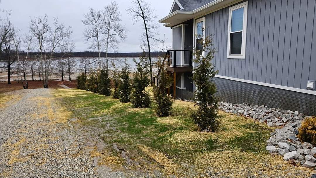 Side view of a gray house with a gravel driveway and small trees lining a patch of grass next to the house.