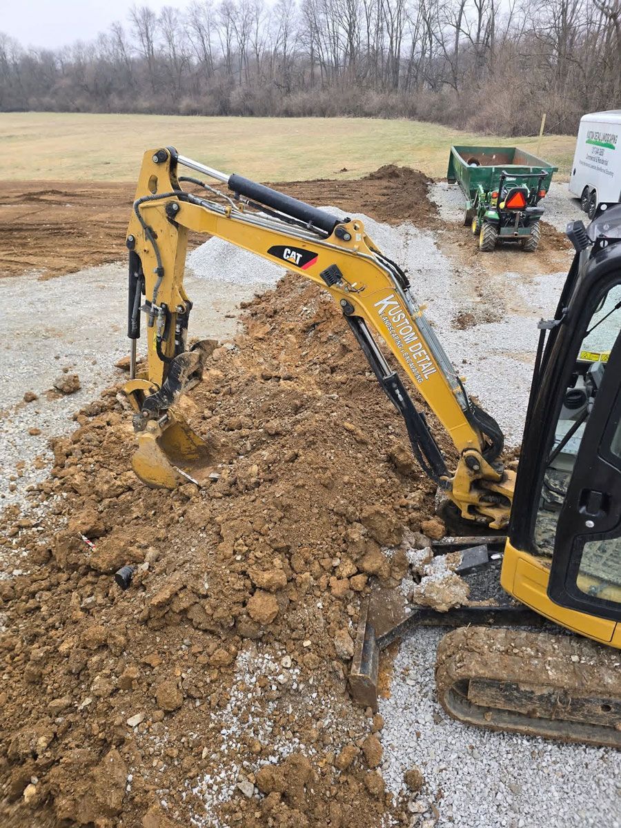 Yellow excavator digging in brown earth, gravel base, green tractor in the background.