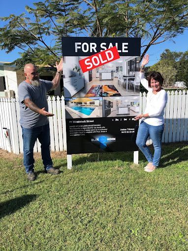 a man and a woman are standing in front of a sold sign after buying a house in Robina