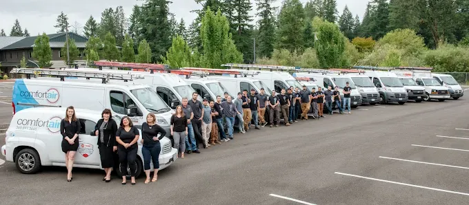 A large group stands in a parking lot in front of a long row of white service vans branded with the Comfort Flow logo.
