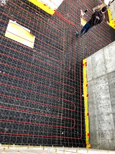 A worker walks across a grid of black flooring panels with red radiant heating tubes installed during construction.