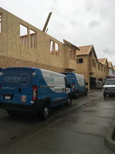 Two blue and white Comfort Air vans parked on a wet street next to multi-story townhomes under construction.