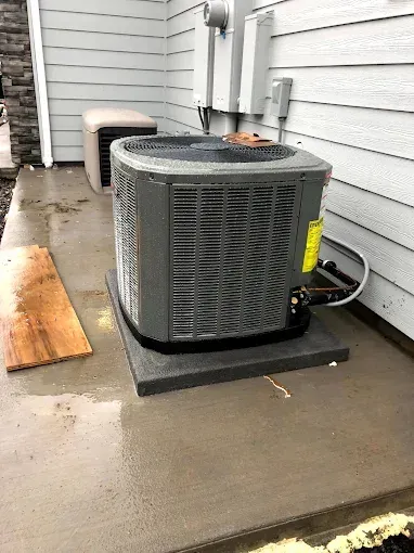 A gray central air conditioning unit sits on a concrete pad next to the light gray siding of a building.