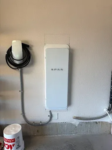 A white Spanner smart electrical panel mounted on a beige wall next to a Tesla wall charger with a coiled charging cable.