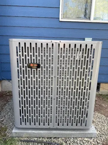A silver Trane air conditioner unit sitting on a gravel base against a blue-sided house.