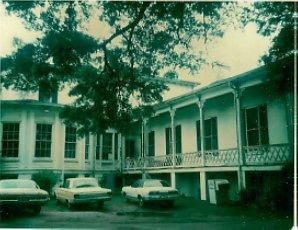 An old white two-story building with a covered balcony, shaded by trees, with three vintage cars parked in the front yard.