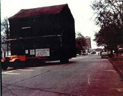 A flatbed truck transports a large, dark-colored wooden building down a quiet paved street.