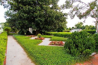 A gravel path winds through a landscaped garden featuring manicured green hedges, patches of red flowers, and a large tree.
