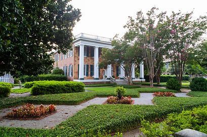 A two-story brick mansion with white columns stands behind a well-manicured garden with low hedges and flower beds.