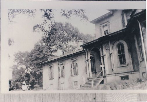 A black-and-white photo shows a historic, multi-story house with a columned porch and arched windows near large trees.