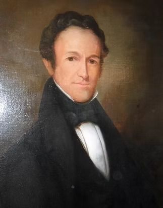 A portrait of a person with wavy dark hair, wearing a high-collared dark coat over a white shirt, against a brown background.