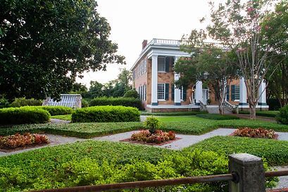 A stately, two-story brick mansion with white columns sits behind a manicured garden with lush hedges and colorful flowers.