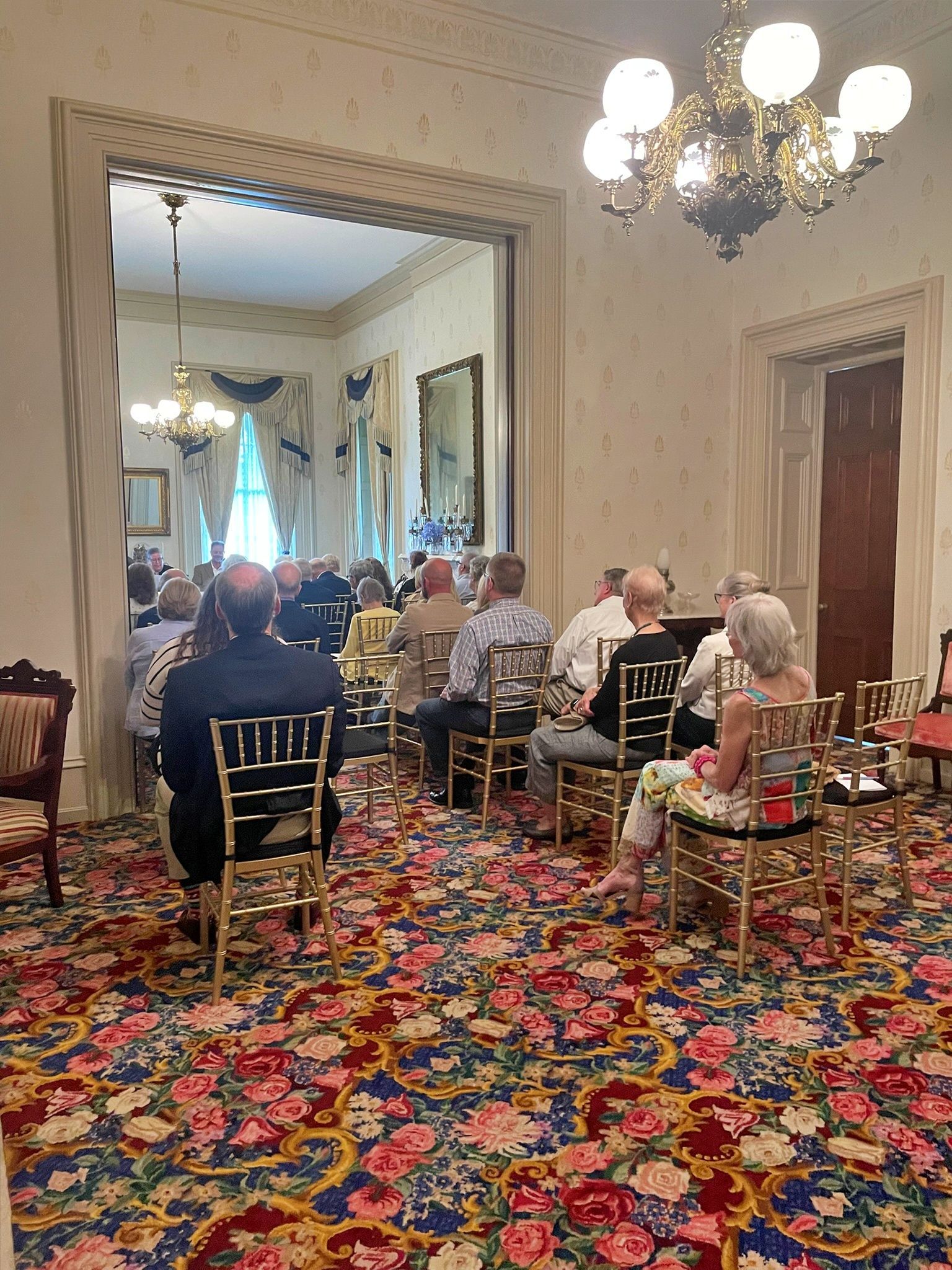 People sit on gold chairs in a room with floral carpeting, attending an event facing a doorway into another lit room.