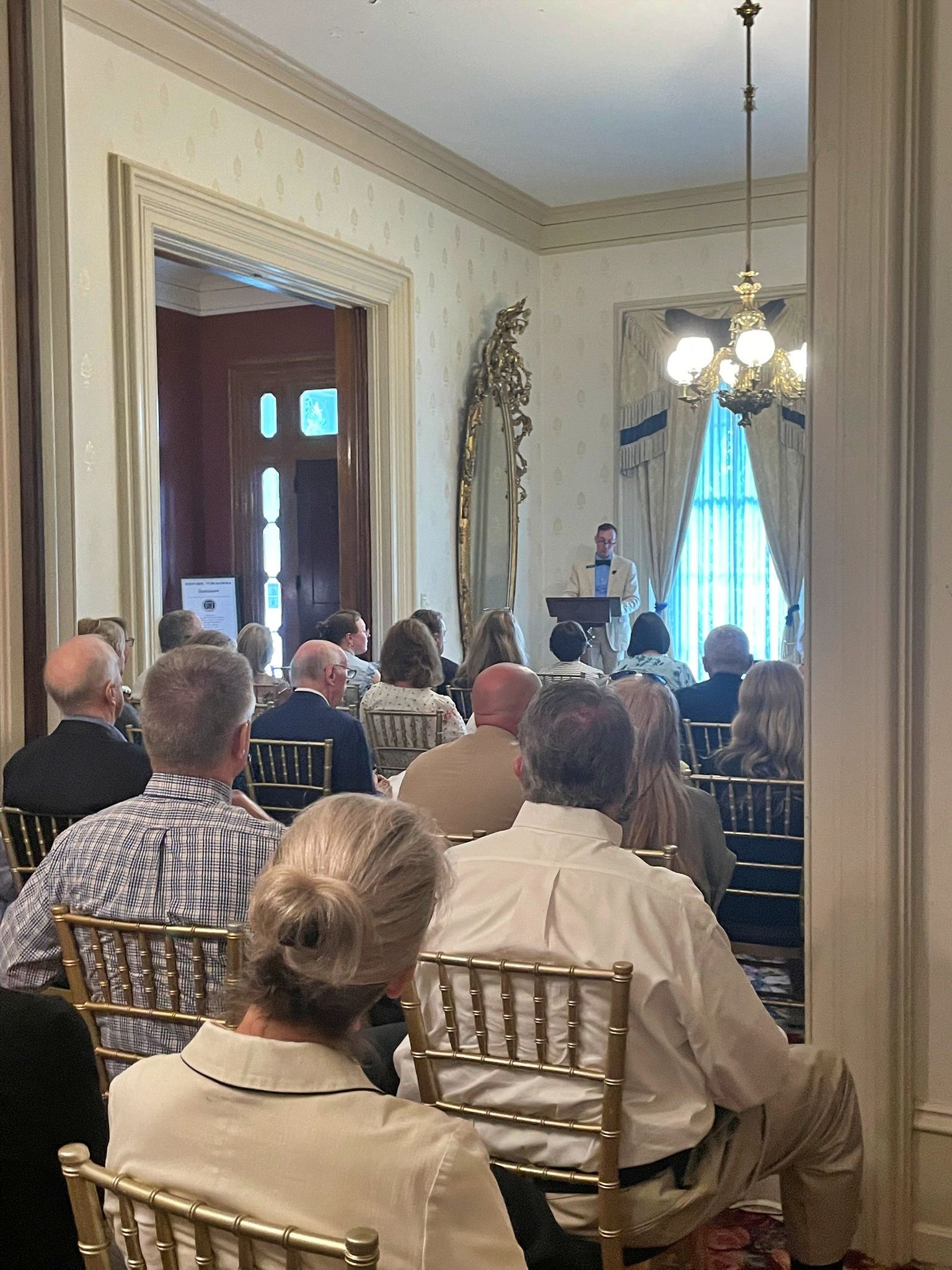 A speaker stands at a podium in a formal, well-lit room, addressing an audience seated in gold chairs.
