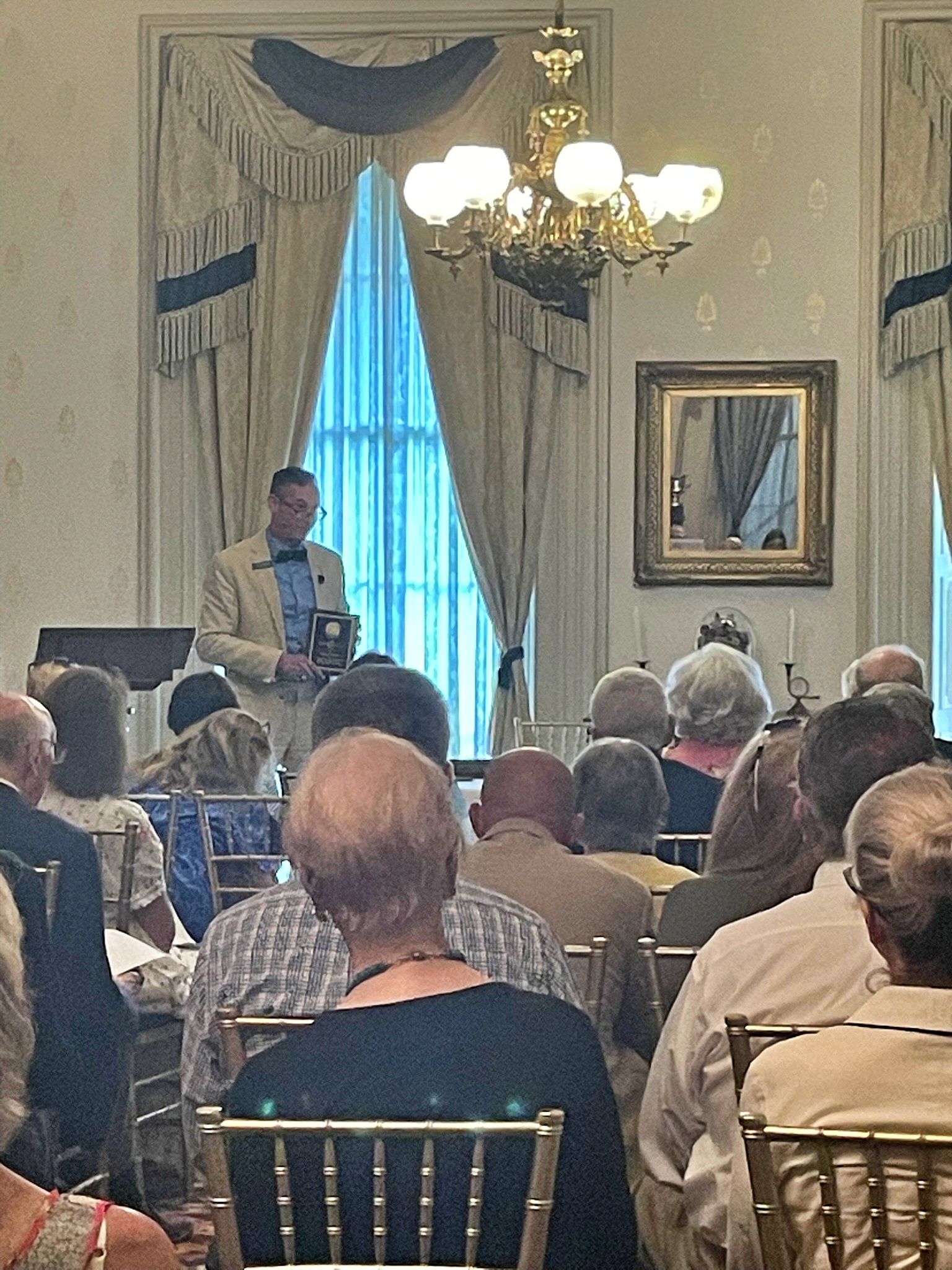 A speaker stands before an audience in an elegant room with tall windows, heavy curtains, and a gold-toned chandelier.