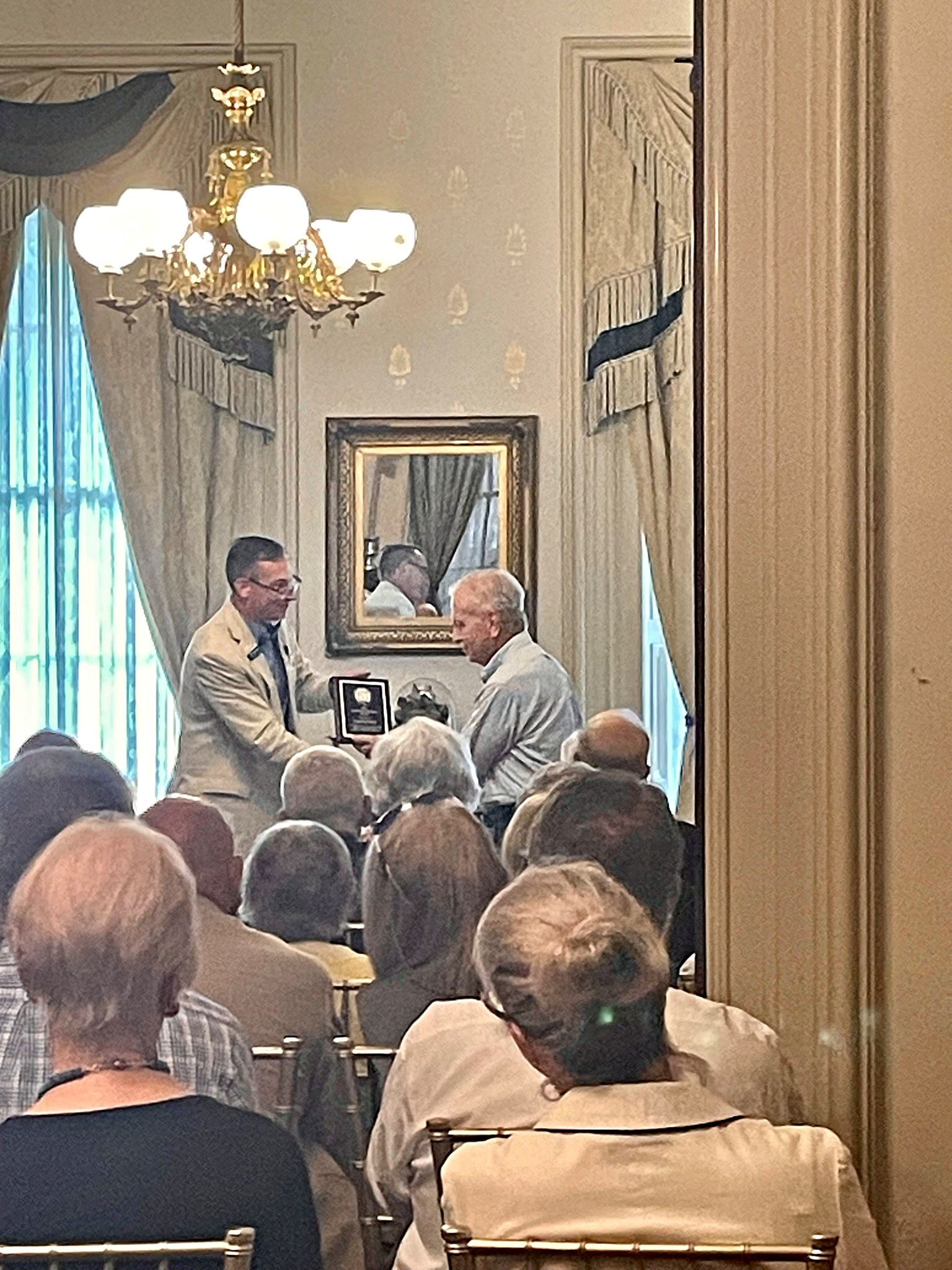 A person in a light suit hands an award to another person in a room with a chandelier, drapery, and framed art.