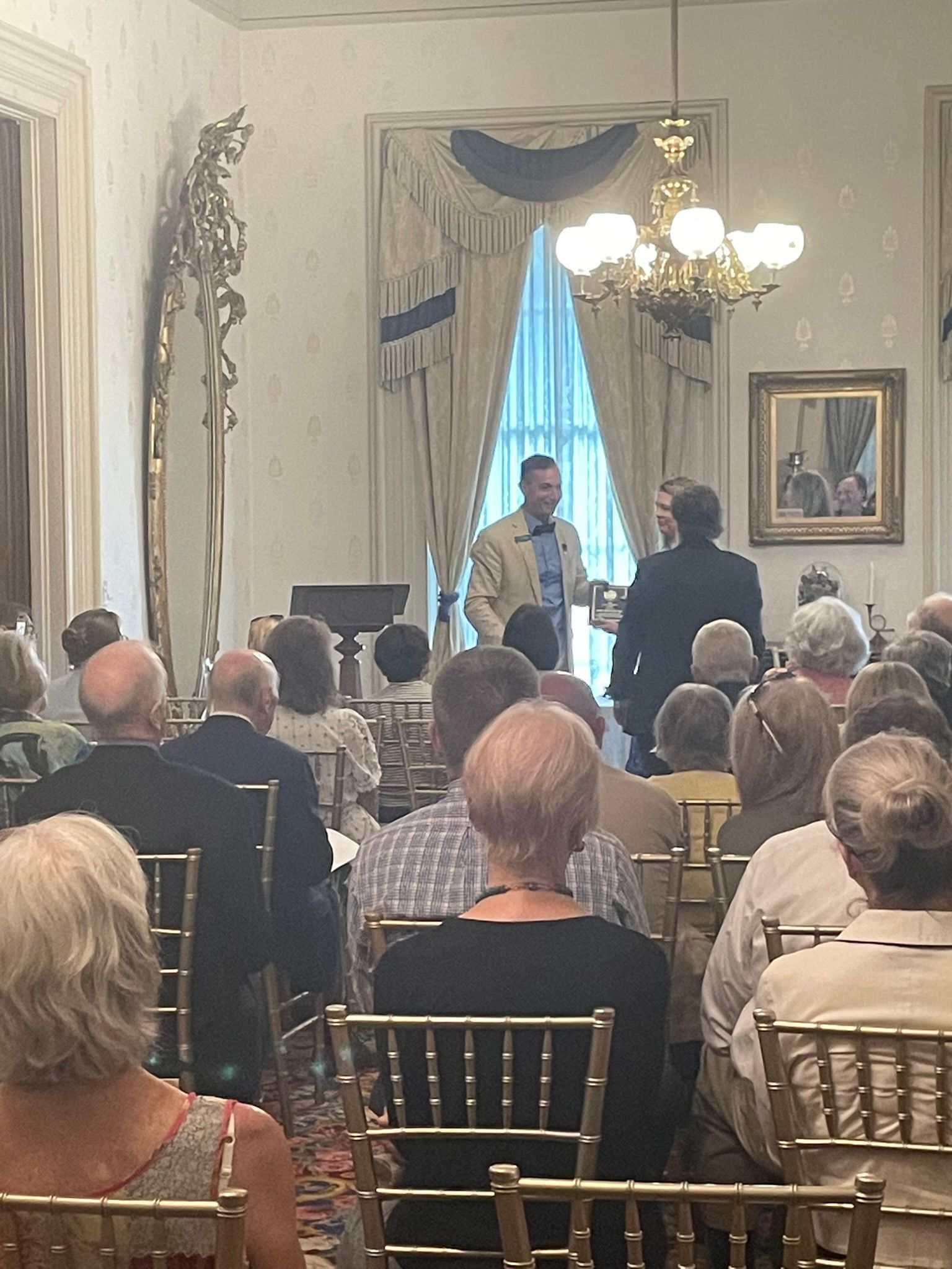 A speaker in a light blazer presents to an audience seated in a formal, well-lit room with tall curtains and a chandelier.