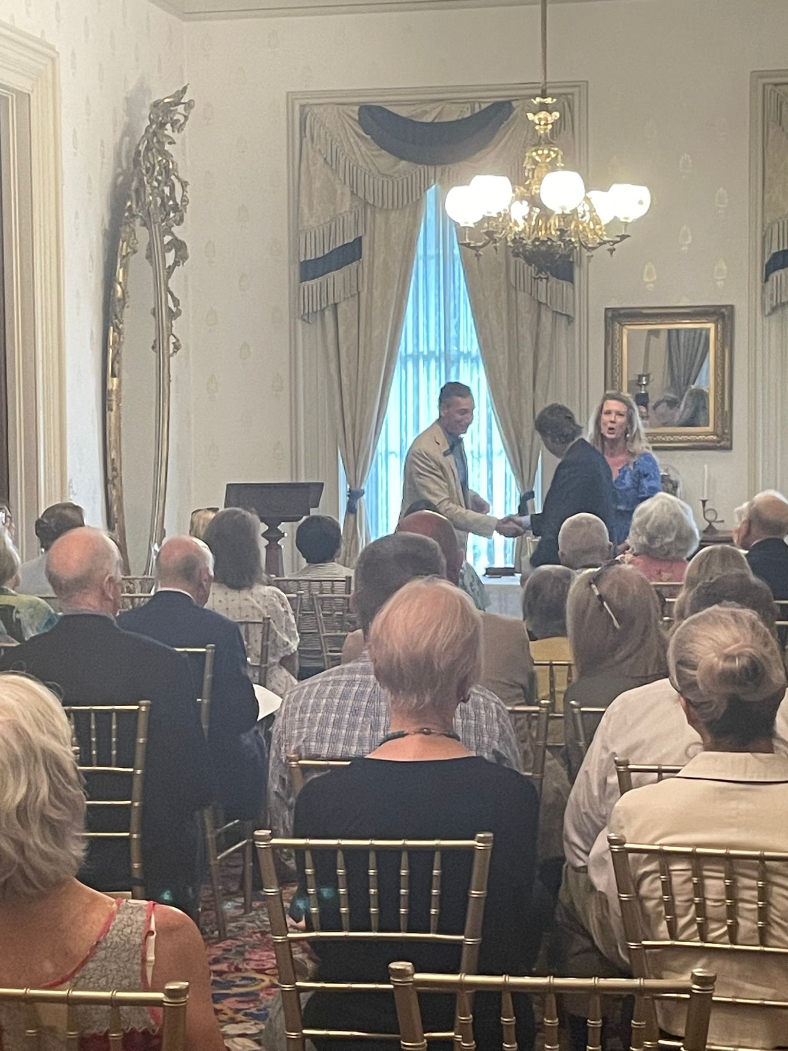 A person in uniform shakes hands with another during an indoor ceremony, as an audience watches from chairs.