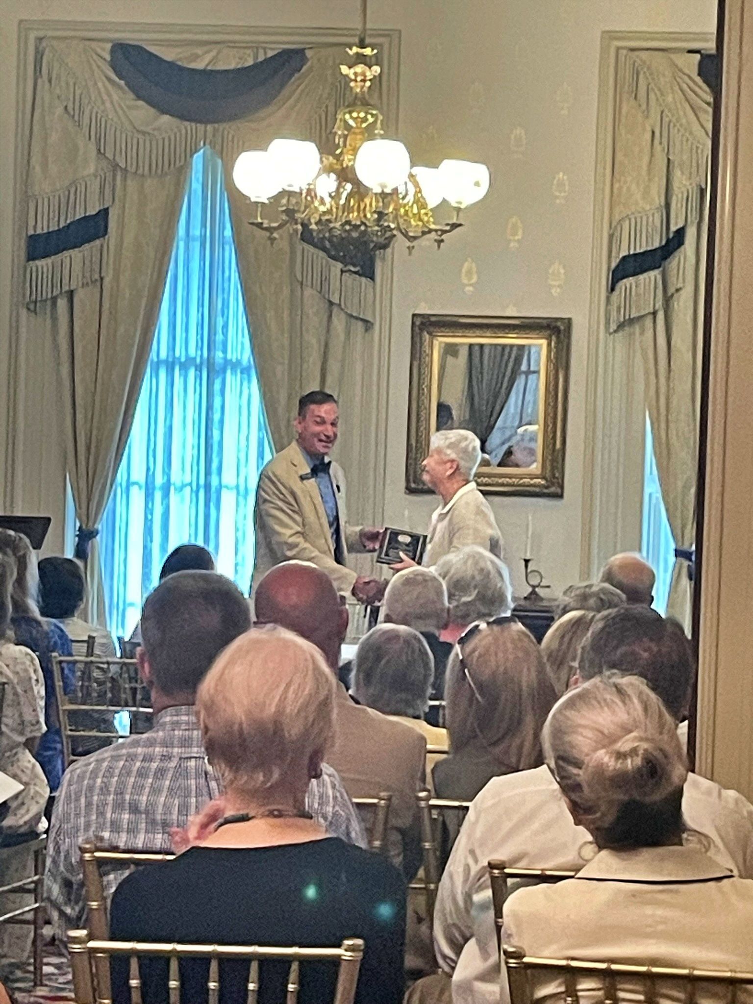 A presenter hands an award to a recipient in a formal room with a chandelier, curtains, and an audience in the foreground.